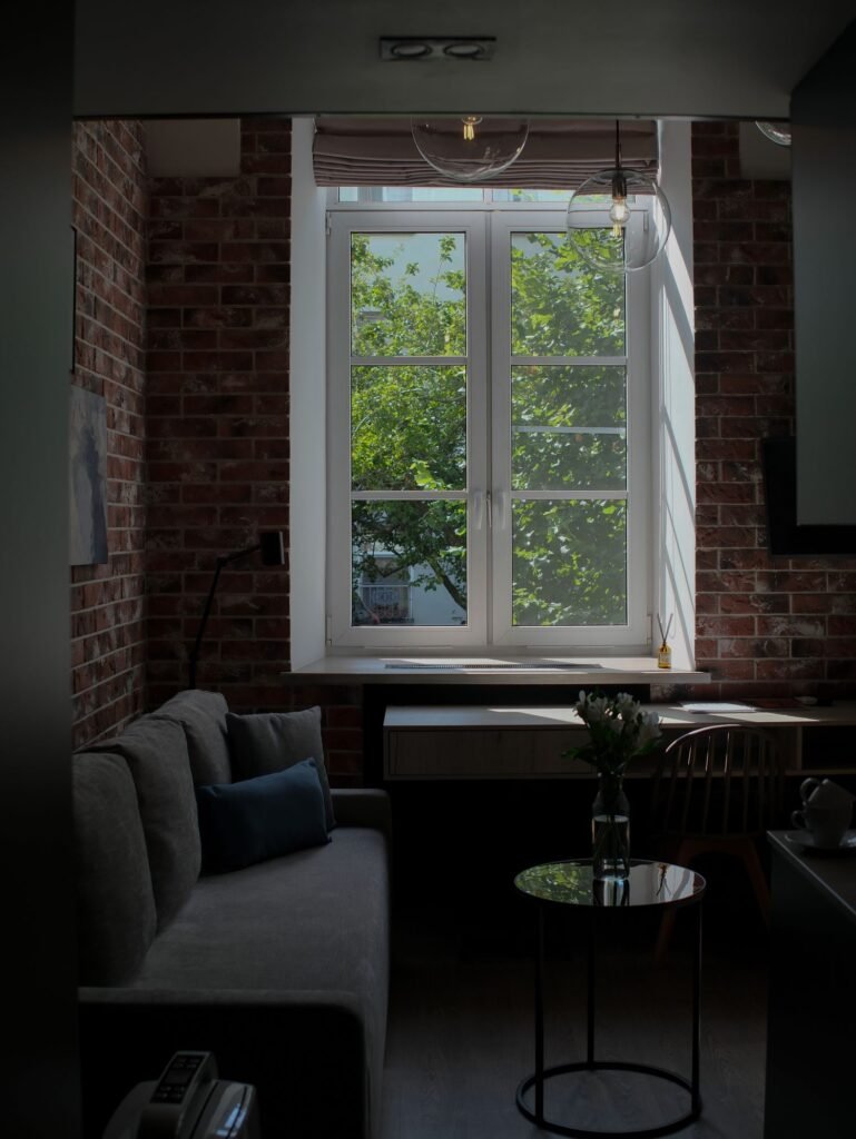 Living room with exposed brick walls, a gray sofa and a small glass side table beside a tall window